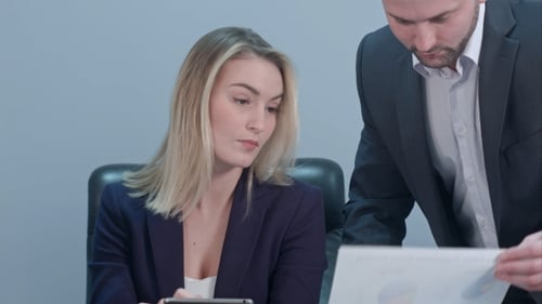 Young Female Boss Sitting at Workplace and Reading Paper with Caucasian Colleague in Office