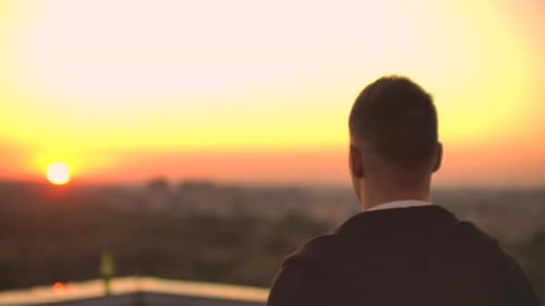 Blick von hinten: Ein Mann geht bei Sonnenuntergang auf dem Dach und trinkt Bier mit Blick auf die schöne Aussicht.