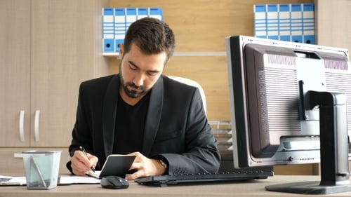 Junger Geschäftsmann in seinem Büro macht sich Notizen in einem Papiernotizbuch