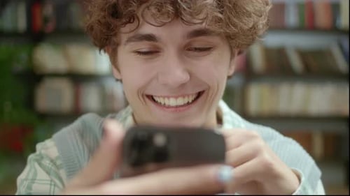 Handsome Curly Student Man Holding Modern Smartphone Texting Message in Home Office or Library