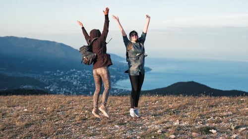 Two Young Happy Women Dancing and Raising Arms on Beautiful Mountain Top Over Sunset Sky