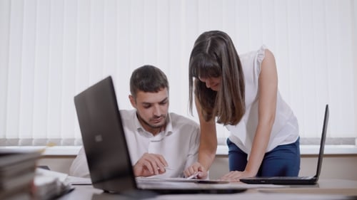 Man Is Sitting on a Chair in Office and Young Woman Is Standing Near, Both Are Looking on a Paper on