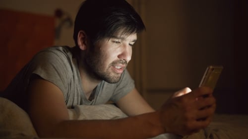 of Young Smiling Man Using Smartphone for Surfing Social Media Lying in Bed at Home at Night