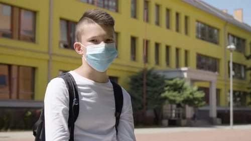A Caucasian Teenage Boy in a Face Mask Looks at the Camera a School in the Background