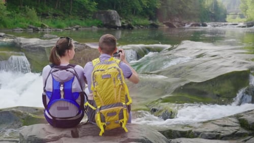 A Young Couple of Tourists Admiring the Mountain River, a Man Taking Pictures of the River with a