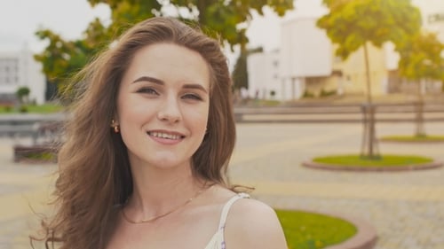 Young Beautiful Girl in Summer Dress in City Park Posing in Front of Camera, Smiling and Adjusting