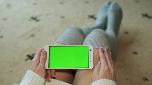 A Woman Is Sitting on the Floor and Holding a Smartphone with a Green Screen