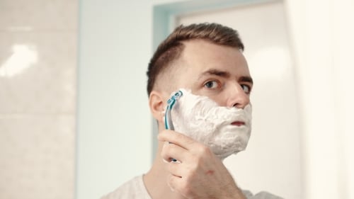 Jovem faz a barba na frente de um espelho no banheiro