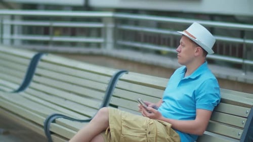 A man in a white hat is resting while sitting on a city bench, tracking a camera