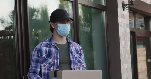 Kurier trägt Maske und Handschuhe mit Paket in der Hand am Gebäude blickt in die Kamera