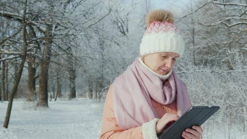 A Woman in a Pink Jacket Enjoys a Walk in a Winter Park. Uses a Digital Tablet