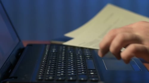 of Man Holding Paper Documents in His Hand and Presses Keys of Laptop at Office