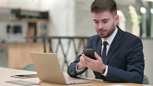 Young Businessman Using Smartphone and Laptop in Office