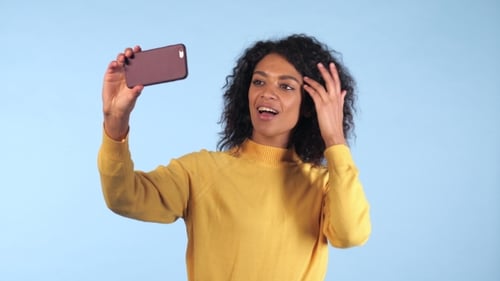 Beautiful African American Woman Holding and Using Smart Phone To Film Herself in Blue Studio. Black