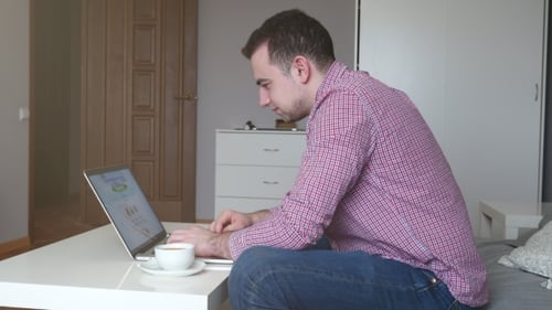Mann arbeitet von zu Hause aus mit Top und Trackpad. Junger Geschäftsmann sitzt am Tisch und trinkt