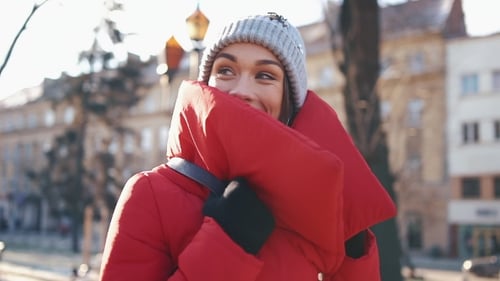 Beautiful Woman in a Red Jacket Stands on the Street Covered with Snow in a Sunny Winter Day