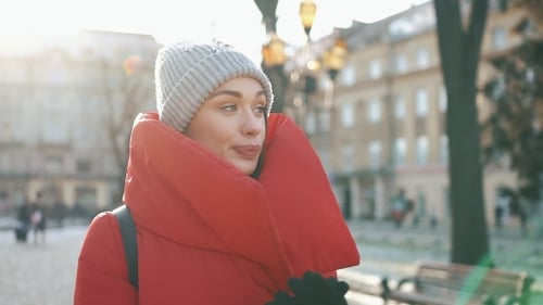 Girl in Grey Hat and Red Coat Walks Along the Street and Looks Around in a Sunny Winter Day