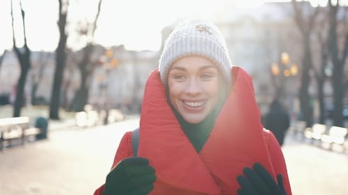 Girl in Grey Hat and Red Coat Walks Along the Street, Smiles and Looks Around in a Sunny Winter Day