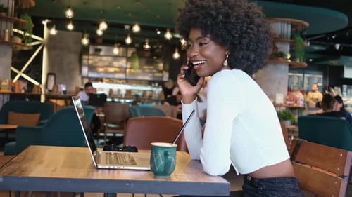 Happy African American Woman Sitting in Cafe with Coffee Talkin on Phone Working on Laptop Freelance