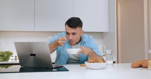 Man Eating Cereal and Using Laptop in Kitchen