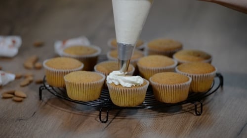 Cream being piped onto cupcakes on a cooling rack
