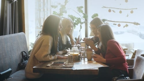 Smiling Girl Showing Something on a Smartphone To Friends While Sitting in a Cafe