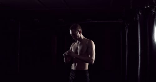 Shirtless Muscular Man Wraps Wrists in Dark Gym