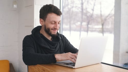 a Young Businessman Working at a Laptop in a Cafe and Smiling