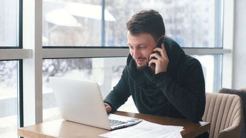 a Young Businessman Talking on the Phone and Working at a Laptop in a Cafe