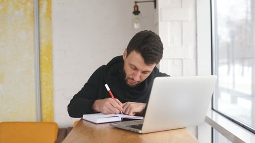 Man Writing in Notebook with Laptop Indoors