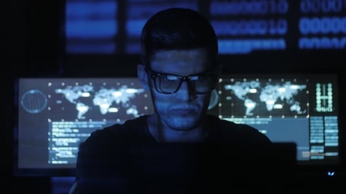 Hacker Programmer in Glasses Is Working on Computer While Blue Binary Code Characters Reflect on His
