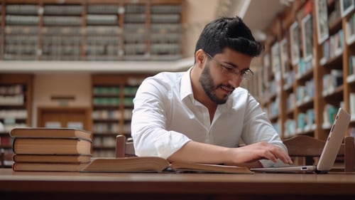 Man Reads Book and Types on Laptop in Library