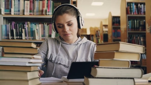 Positive junge lächelnde Frau in weißen Kopfhörern sitzt am Tisch in der Universitätsbibliothek