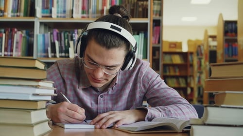 Young Caucasian Male Student with Big Headphones Is Sitting at Table in Library. Many Books Are