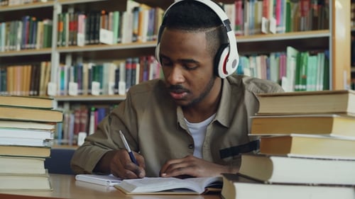 Hübscher afroamerikanischer Student, der Kopfhörer trägt, hört Musik und sitzt am Tisch in
