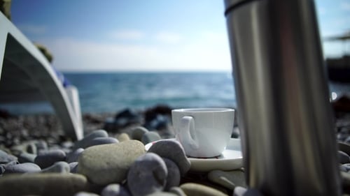 A Cup of Coffee with Blue Sea. White Cup on the Rocks on the Shore