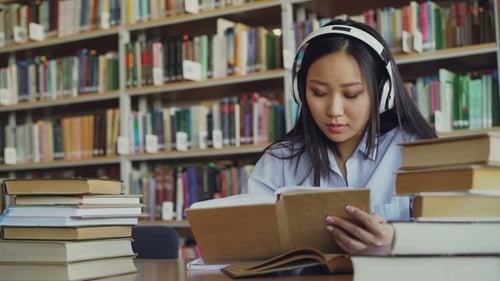 Une étudiante asiatique assez positive avec des écouteurs sur la tête écoutant de la musique est assise à la table