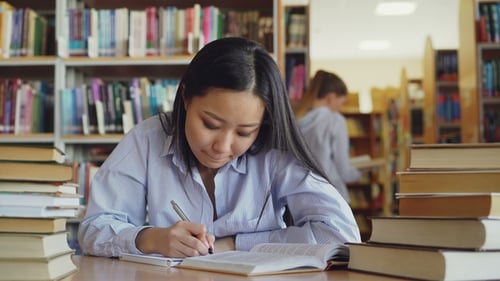 Young Beautiful Asian Female Student Is Sitting at Table Surrounded By Piles of Books in Big Library