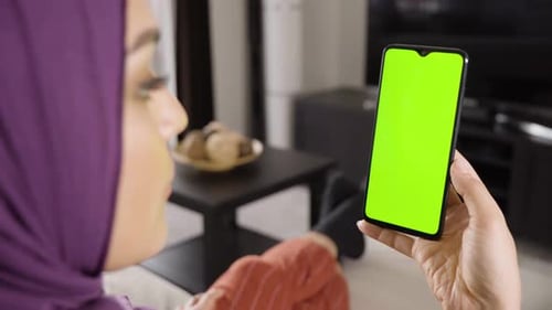 A Muslim Woman Looks at a Smartphone with Green Screen in an Apartment Closeup From Behind
