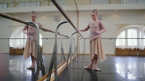 Ballerina in Beige Sweatsuit and Pointe Stretches on Barre in Ballet Gym. Woman Standing Near Bar