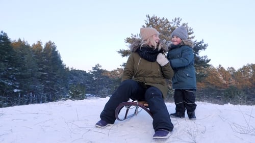 Little Boy Pushes Mother on Sledge and She Rides from Snowy Hill in Winter Forest