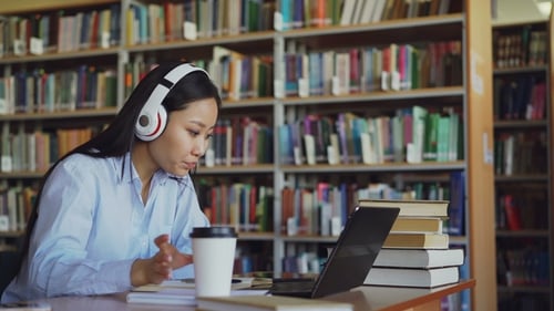 Beautiful Asian Female Student Listening Music in Headphones Sitting at Desk in Library Working at