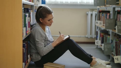 Junge schöne verträumte Studentin sitzt auf dem Boden in der leichten Bibliothek zwischen Bücherregalen und schreibt