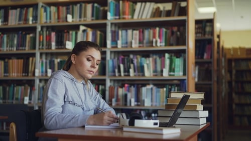 Beautiful Serious Caucasian Female Student Sitting at Table with Books in Library Writing Down