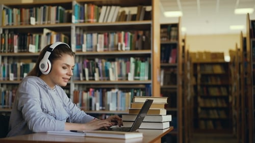 Beautiful Positive Caucasian Female Student with Big Headphones Working at Table in Spacious Library