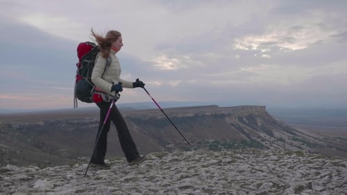 Young Hiker Woman with Backpack and Hiking Poles Is Walking Along the Edge of Rock