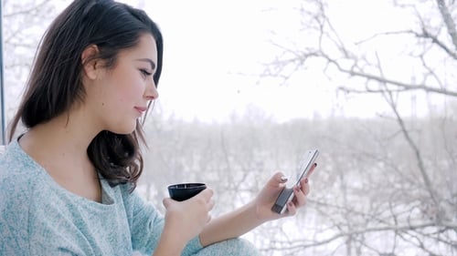 Young Woman Drinking Coffee With Phone By Window