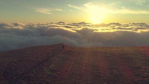 Young Woman Is Walking Above Pink Clouds to the Edge of Mountain Plateau at Sunrise