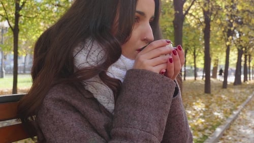 Young Southern Brunette Woman in Grey Coat Is Drinking Hot Beverage from Thermos Mug in Autumn Park