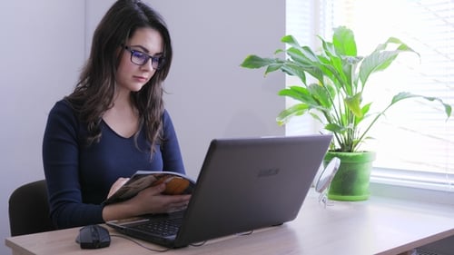 Businesswoman in Eyeglasses Flipping Magazine Sitting in Front of Laptop Computer at Work